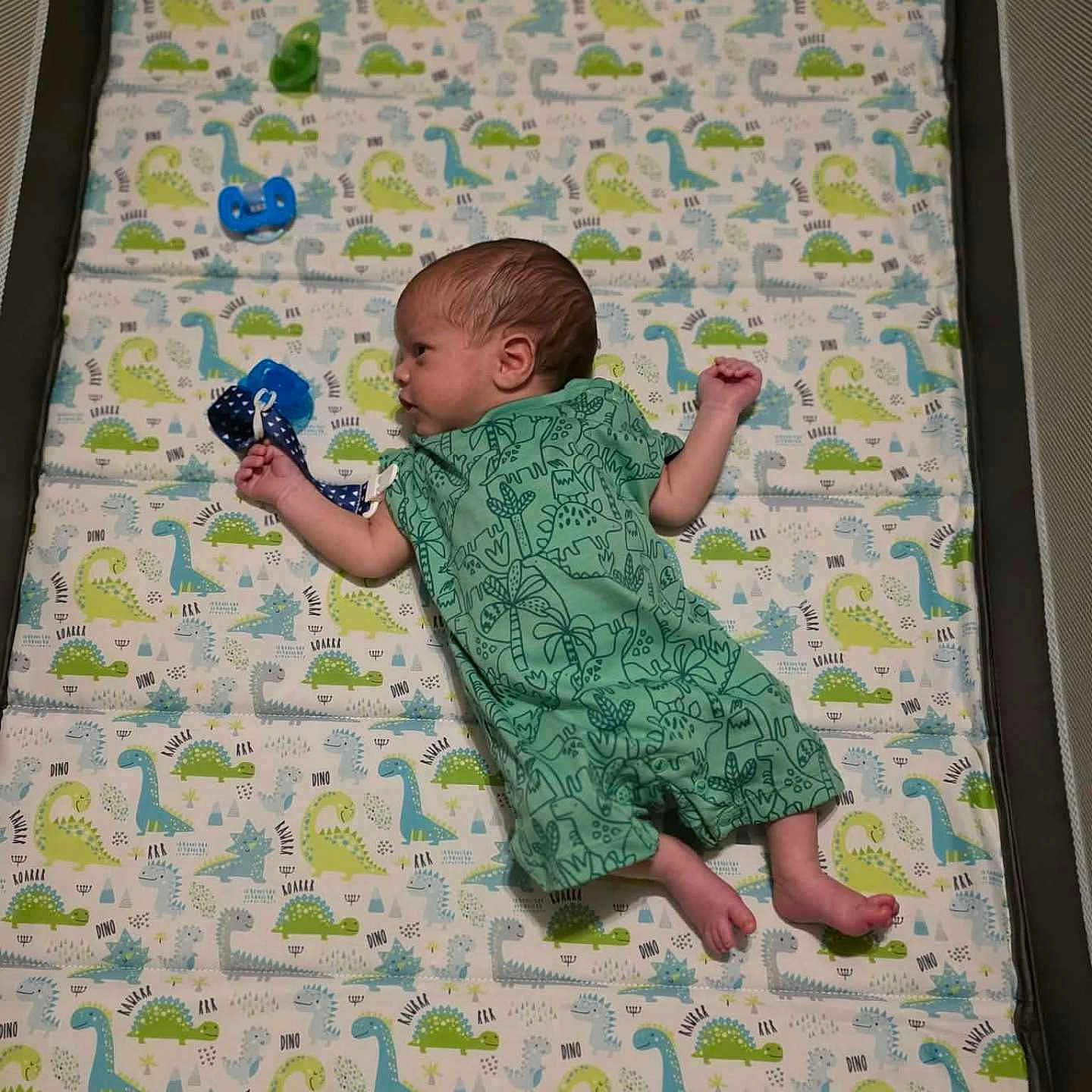 Daniel is registered to the contest to win money with this photo: baby, newborn, infant, child, lying_down, changing_mat, dinosaur_pattern, green_outfit, onesie, toy, blue_toy, portrait, side_profile, indoor, nursery, baby_gear, sleeping_area, feet, playful, family_photo