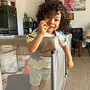 Nolan a rejoint le concours — aidez-le/la à gagner de superbes lots ! toddler, child, curly_hair, playpen, indoor, smiling, kitchen, dining_room, furniture, painting, table, chair, floor, tile_floor, cabinet, appliance, toy, colorful, happy, person