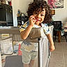 toddler, child, curly_hair, playpen, indoor, smiling, kitchen, dining_room, furniture, painting, table, chair, floor, tile_floor, cabinet, appliance, toy, colorful, happy, person