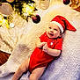 baby, infant, christmas, holiday, red_clothing, santa_hat, smile, blanket, tree, ornaments, lights, festive, indoors, cute, happy, lying_down, celebration, decoration, seasonal, person