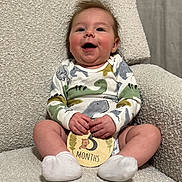 Archer is registered to the contest to win money with this photo: baby, child, infant, sitting, smiling, onesie, dinosaur_pattern, milestone_card, white_socks, armchair, beige, texture, hands, face, happy, cute, indoor, portrait, young, cozy