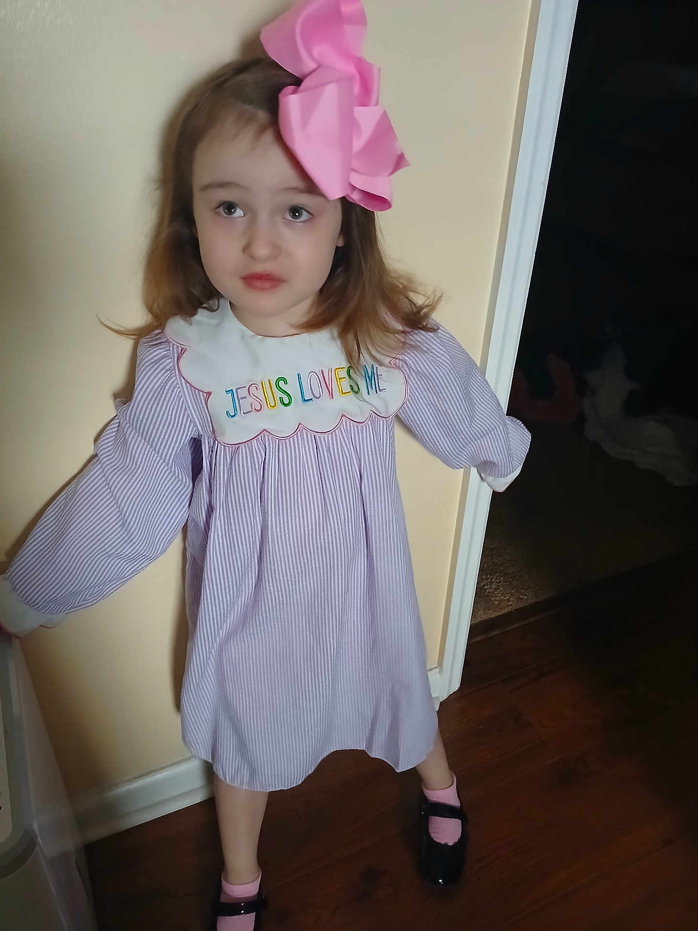 Adriana is registered to the contest to win money with this photo: child, girl, hair_bow, pink_bow, striped_dress, embroidered_text, peter_pan_collar, indoor, doorway, hardwood_floor, pink_socks, black_shoes, mary_janes, standing, portrait, curious_expression, long_hair, home_interior, soft_lighting, wall