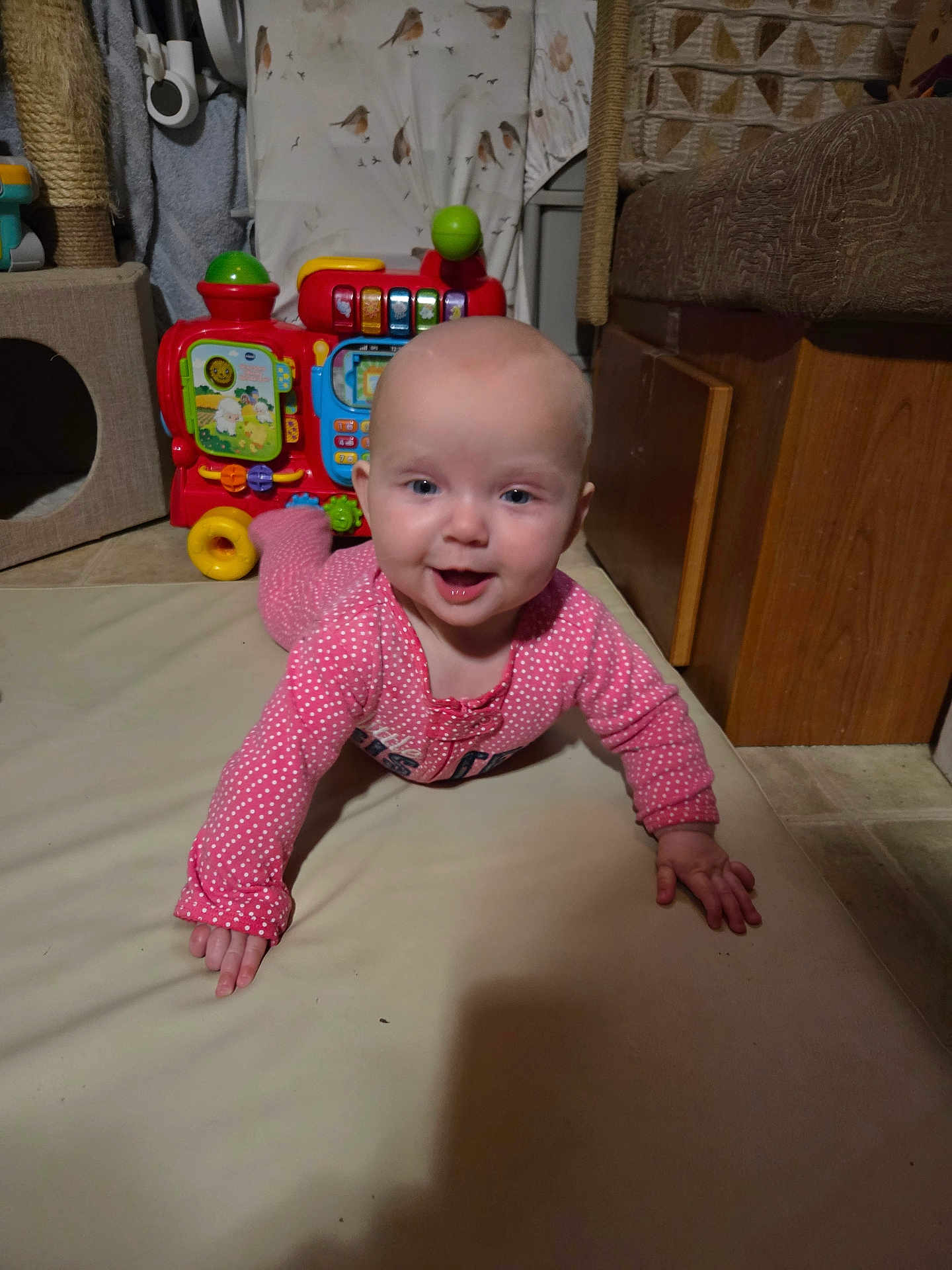 Lola joined the competition — help win amazing prizes! baby, infant, crawling, pink_onesie, toy, indoor, floor_mat, happy, child, smiling, face, head, hand, wooden_furniture, colorful, playtime, cute, home, floor, expression