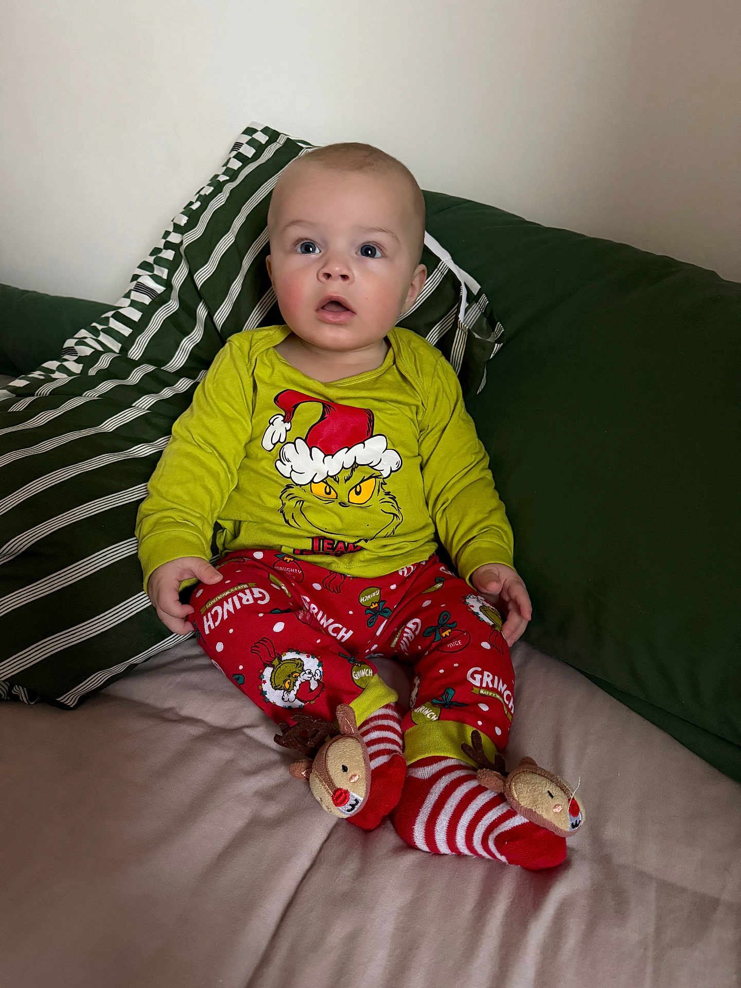 Jesse is registered to the contest to win money with this photo: baby, child, pajamas, grinch, socks, reindeer, bed, pillow, green, striped, clothing, person, indoor, cute, portrait, seated, expression, holiday, festive, comfortable