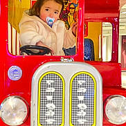 Harvy a rejoint le concours — aidez-le/la à gagner de superbes lots ! toddler, child, pacifier, toy_bus, red, beige_jacket, indoor, steering_wheel, play_area, colorful, face, portrait, cute, window, cartoon, seat, person, playground, clothing, young_child