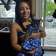 Willow is registered to the contest to win money with this photo: woman, person, cat, gray_cat, pet, smiling, braces, portrait, indoor, kitchen, window, daylight, blue_clothing, floral_pattern, daisy, holding_pet, arms, sink, deck, fashionable_pet