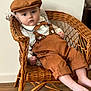 adorable, baby, bow_tie, brown_clothing, cap, casual_pose, child, curious, cute, fashion, furniture, indoors, infant, looking_at_camera, portrait, seated, soft_lighting, suspenders, wicker_chair, wooden_floor