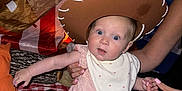 Isla is registered to the contest to win money with this photo: adult, baby, bib, blue_mat, bright_eyes, child, clothing, cowboy_hat, curious, face, golden_ball, hand, indoor, party_decoration, person, pillow, pink_outfit, seated, soft_texture, teepee