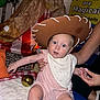 adult, baby, bib, blue_mat, bright_eyes, child, clothing, cowboy_hat, curious, face, golden_ball, hand, indoor, party_decoration, person, pillow, pink_outfit, seated, soft_texture, teepee