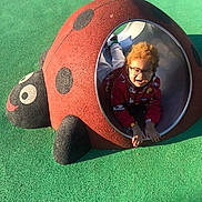 Noha participe au concours pour gagner de l'argent avec cette photo : black_spots, casual_clothing, child, crawling, curly_hair, daytime, fun, glasses, green_floor, happy, joy, kids_play_area, ladybug, outdoor, playground, recreation, red, smiling, sunlight, tunnel