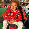 casual_clothing, child, curly_hair, daylight, glasses, green_surface, mickey_mouse, outdoor, park, people_in_background, playground, red_jacket, serious_expression, sitting, smiling_toy, sneakers, sunlight, toy, trees, white_pants