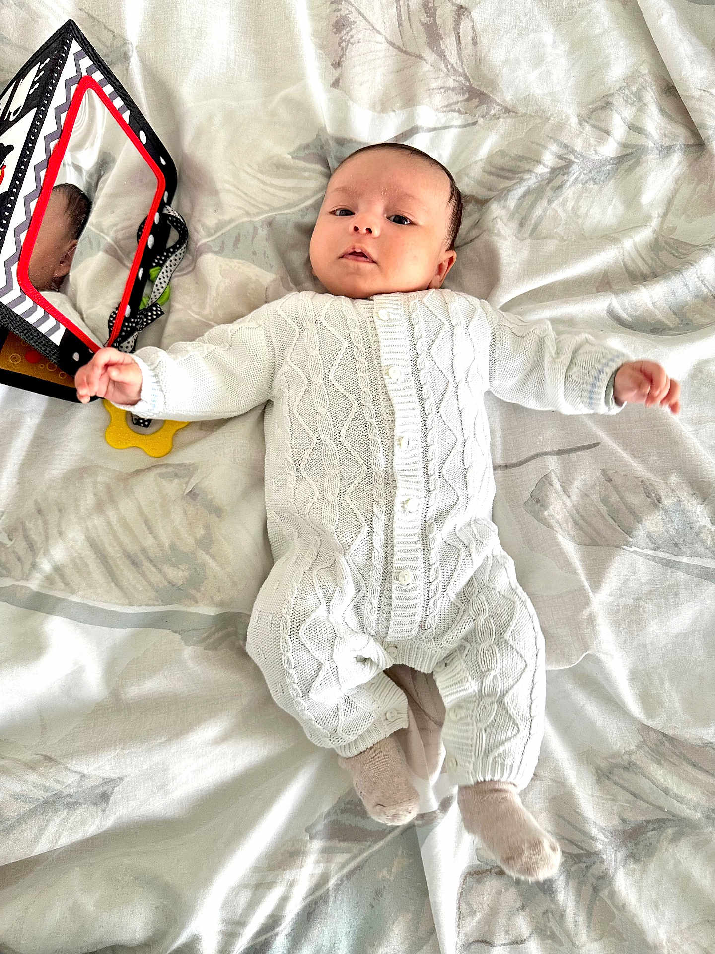 Iyad a rejoint le concours — aidez-le/la à gagner de superbes lots ! baby, infant, white_knitted_outfit, blanket, toy, mirror, reflection, soft_lighting, lying_down, curious, indoor, child, cute, patterned_blanket, socks, small_hands, face, head, comfortable, peaceful