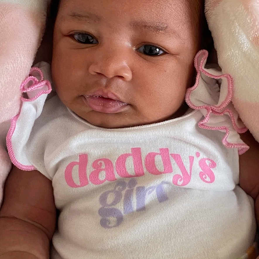 Audree joined the competition — help win amazing prizes! baby, infant, child, portrait, lying_down, blanket, bow, hair_accessory, onesie, striped_clothing, rainbow_colors, pastel, brown_skin, dark_hair, wide_eyes, serious_expression, indoor, close_up, text_on_clothing, family_theme
