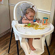Violet is registered to the contest to win money with this photo: toddler, highchair, food, messy, baby, spoon, sippy_cup, floor, wooden_floor, child, indoor, feeding, concentration, plastic, clothing, hair_tie, wall, appliance, kitchen, playful
