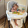 toddler, highchair, food, messy, baby, spoon, sippy_cup, floor, wooden_floor, child, indoor, feeding, concentration, plastic, clothing, hair_tie, wall, appliance, kitchen, playful