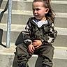 child, toddler, sitting, steps, concrete, camo_jacket, pants, nike_sneakers, shoes, hair_tied, outdoor, sunlight, casual_clothing, person, fashion, street_style, young_child, portrait, curious, daylight
