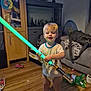 Greyson is registered to the contest to win money with this photo: child, toddler, toy, lightsaber, indoor, living_room, floor, wood_floor, socks, couch, bag, cabinet, toy_car, play_mat, smiling, standing, blond_hair, face, furniture, home