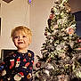 child, toddler, christmas_tree, holiday, festive_clothing, pajamas, blond_hair, indoors, decorations, ornaments, lights, silver_tinsel, smiling, happy, young_child, celebration, seasonal, home, festivity, cute