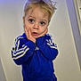 child, blonde_hair, blue_eyes, scar, blue_sweatshirt, adidas, hands_on_face, expression, indoor, casual_clothing, portrait, young_child, curly_hair, standing, light_background, surprised, cute, sportswear, long_sleeves, face