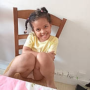 Safiatou a rejoint le concours — aidez-le/la à gagner de superbes lots ! child, smiling, braided_hair, yellow_shirt, wooden_chair, indoor, tablecloth, pink, white_wall, electrical_outlets, knees, happy, seated, casual, portrait, cute, girl, room, furniture, home