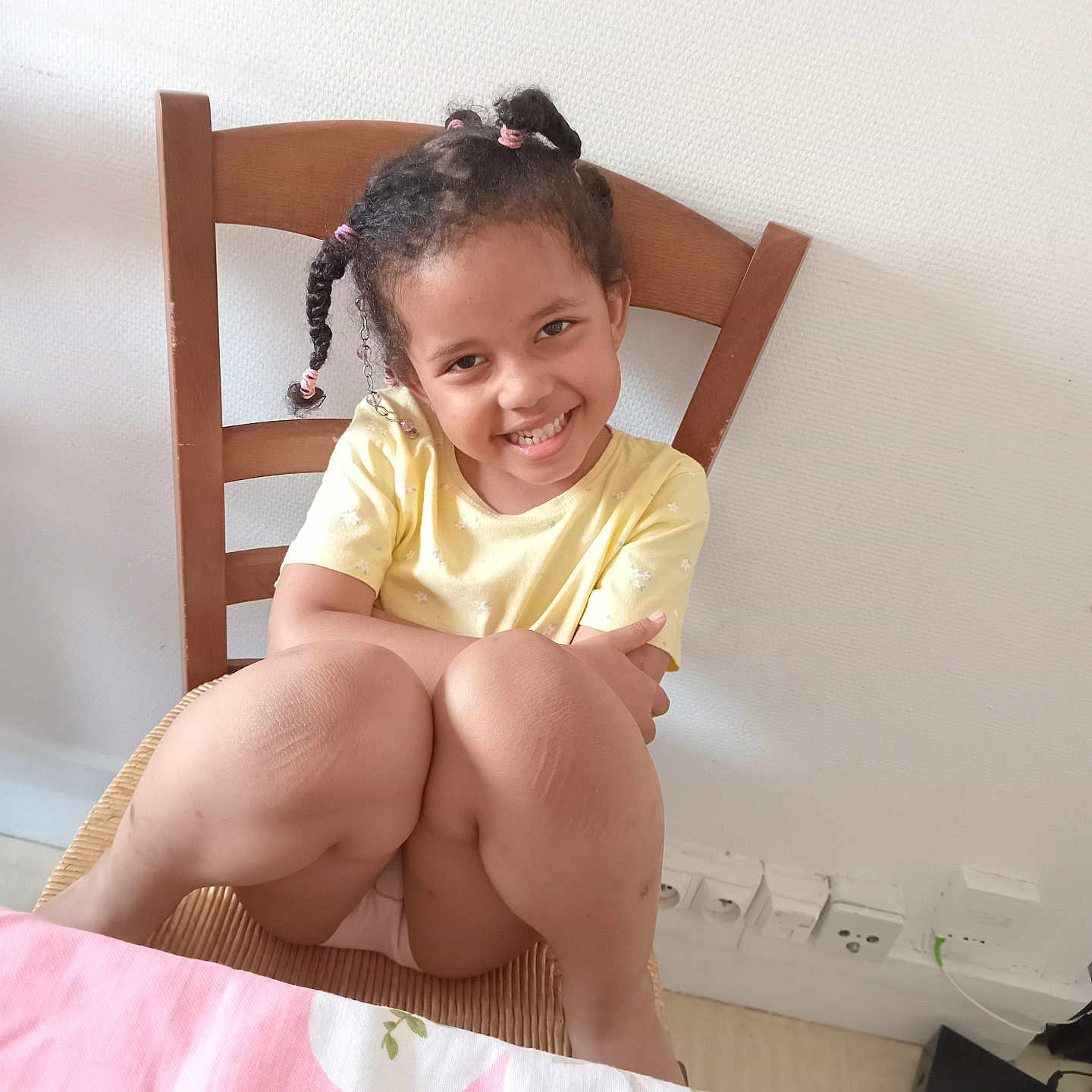 Safiatou a rejoint le concours — aidez-le/la à gagner de superbes lots ! braided_hair, casual, child, cute, electrical_outlets, furniture, girl, happy, home, indoor, knees, pink, portrait, room, seated, smiling, tablecloth, white_wall, wooden_chair, yellow_shirt