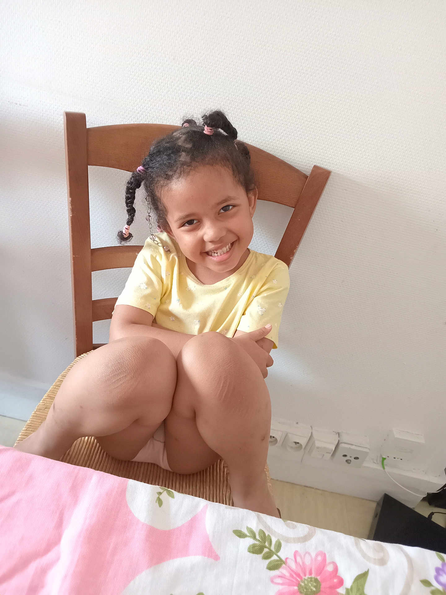 Safiatou a rejoint le concours — aidez-le/la à gagner de superbes lots ! child, smiling, braided_hair, yellow_shirt, wooden_chair, indoor, tablecloth, pink, white_wall, electrical_outlets, knees, happy, seated, casual, portrait, cute, girl, room, furniture, home