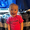 blanket, bright_color, checkered_pattern, child, clothing, curtain, cute, expression, face, happy, indoor, joy, person, pink_shirt, portrait, seated, smiling, text_on_clothing, toddler, young_child