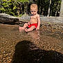 toddler, child, water, red_shorts, nature, forest, pebbles, sunlight, shadow, exploring, summer, outdoor, barefoot, sitting, curious, log, reflection, daylight, skin, riverbank