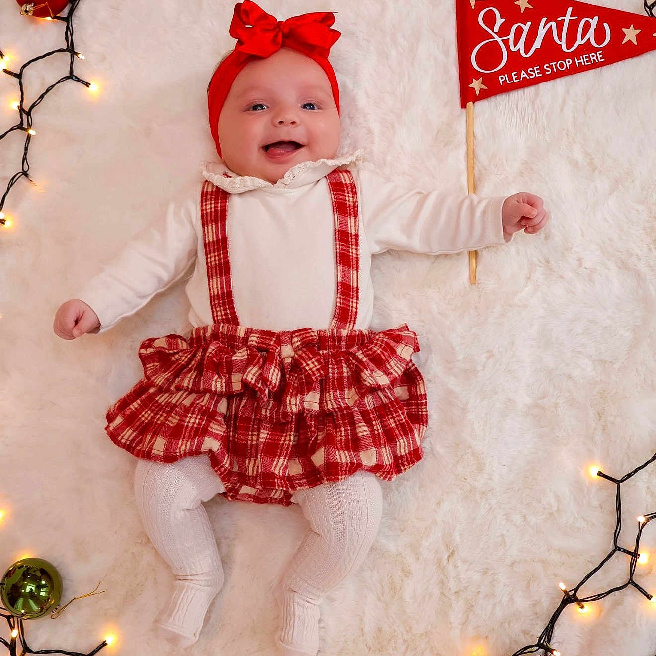 Renesmé is registered to the contest to win money with this photo: baby, blanket, celebration, child, christmas_lights, cute, festive, happy, holiday, infant, lying_down, ornaments, plaid_skirt, red, red_headband, seasonal, sign, smiling, white, white_tights