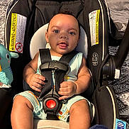 Kayden is registered to the contest to win money with this photo: baby, car_seat, child, infant, smiling, blue_sandals, romper, safety_harness, plush_toy, indoor, bedspread, patterned, cute, happy, person, seat, head, hands, footwear, portrait