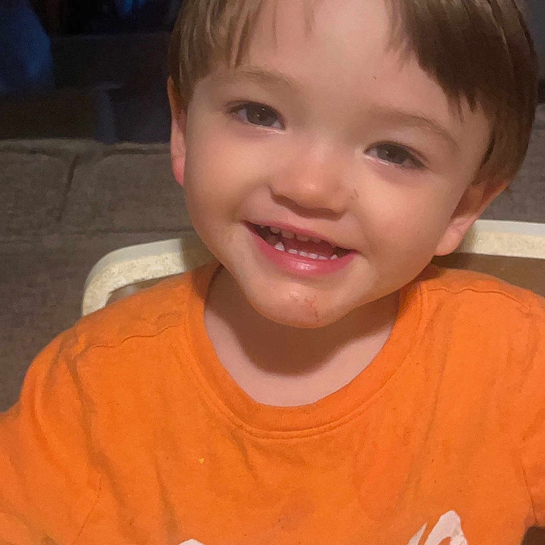 Adam is registered to the contest to win money with this photo: baby, brown_hair, casual_clothing, child, childhood, cute, expression, face, happy, high_chair, home, indoor, messy_shirt, orange_shirt, person, portrait, seated, smiling, toddler, young_child
