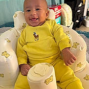 Zion is registered to the contest to win money with this photo: baby, child, smiling, yellow_clothing, indoor, cushioned_chair, teddy_bear_pattern, happy, infant, person, furniture, home, cute, portrait, seated, cozy, soft_lighting, hand, foot, socks