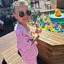 child, girl, sunglasses, pink_clothing, rubber_duck, colorful, outdoor, sunlight, smile, toy_pool, wooden_structure, summer, happy, feet, casual_shoes, playful, person, hair_bun, accessory, recreation