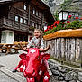 child, girl, statue, red_cow, mountain, wooden_building, flowers, outdoor, bench, pavement, rocks, lantern, smiling, playful, nature, scenic, casual_clothing, daylight, tourist_spot, happy