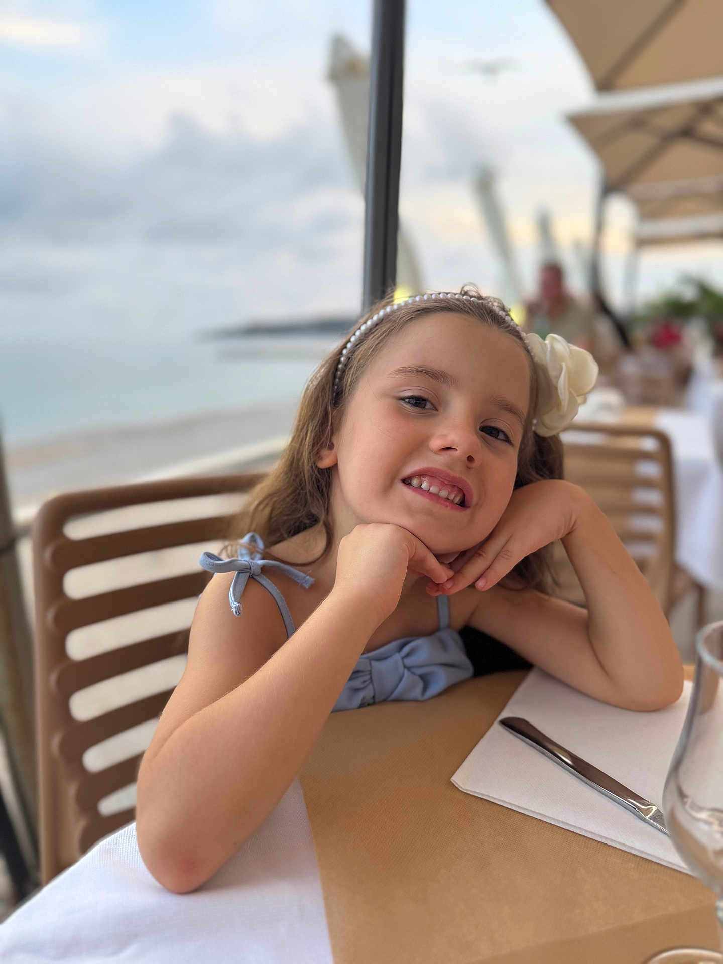 Varvara Bachmaha participe au concours pour gagner de l'argent avec cette photo : child, girl, smile, portrait, headband, flower, restaurant, table, knife, napkin, glass, chair, outdoor, beach, sea, sky, clouds, dining, happy, casual