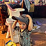 toddler, child, hat, onesie, indoor, living_room, high_chair, laundry_basket, floor, clothing, person, wooden_floor, toy, household_items, cap, baby, furniture, carpet, messy_room, cute