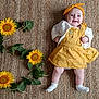baby, child, smiling, yellow_dress, headband, sunflowers, green_leaves, woven_rug, indoor, laying_down, happy, cute, infant, white_socks, natural_light, portrait, flower_arrangement, celebration, number_three, skin