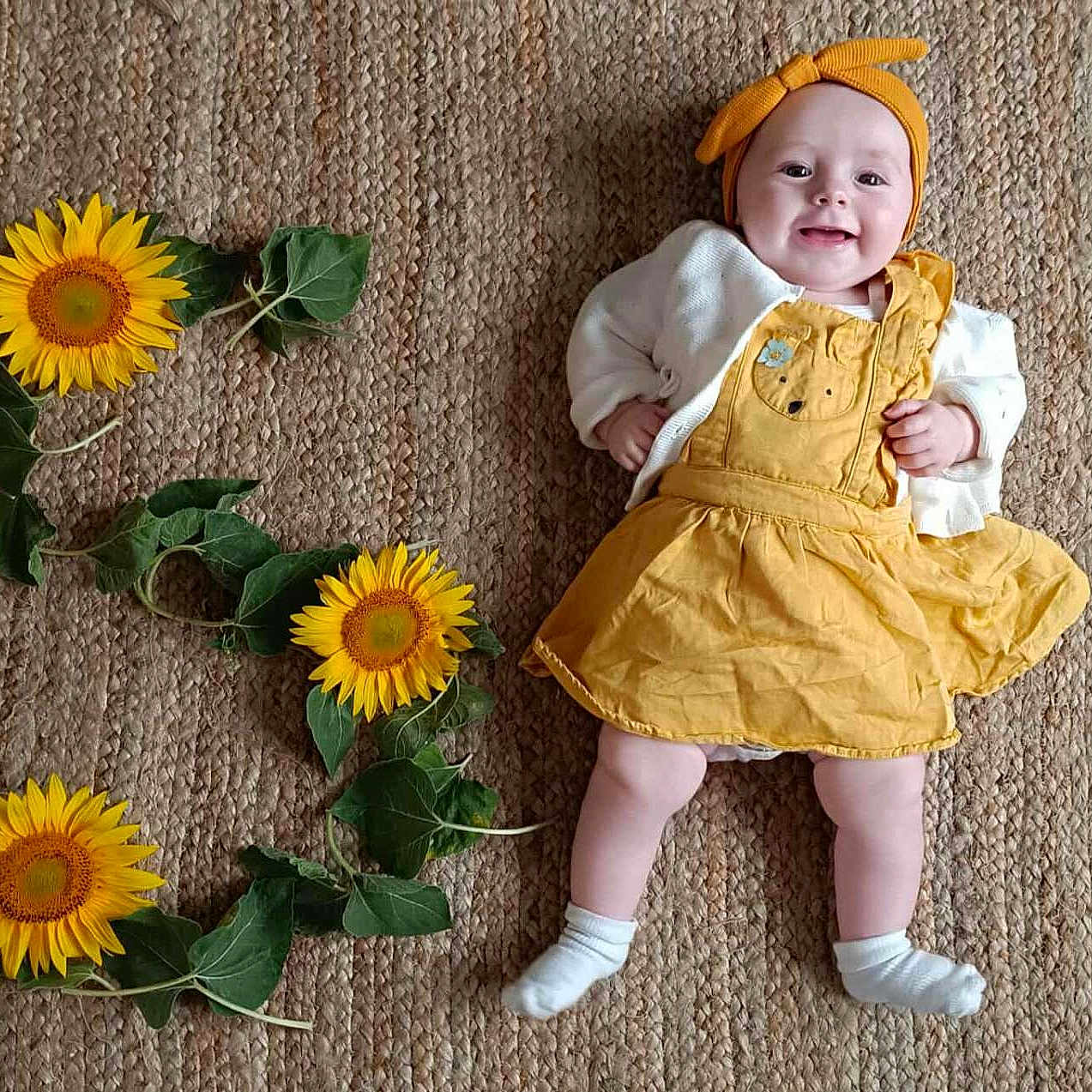 Rosalya participe au concours pour gagner de l'argent avec cette photo : baby, celebration, child, cute, flower_arrangement, green_leaves, happy, headband, indoor, infant, laying_down, natural_light, number_three, portrait, skin, smiling, sunflowers, white_socks, woven_rug, yellow_dress