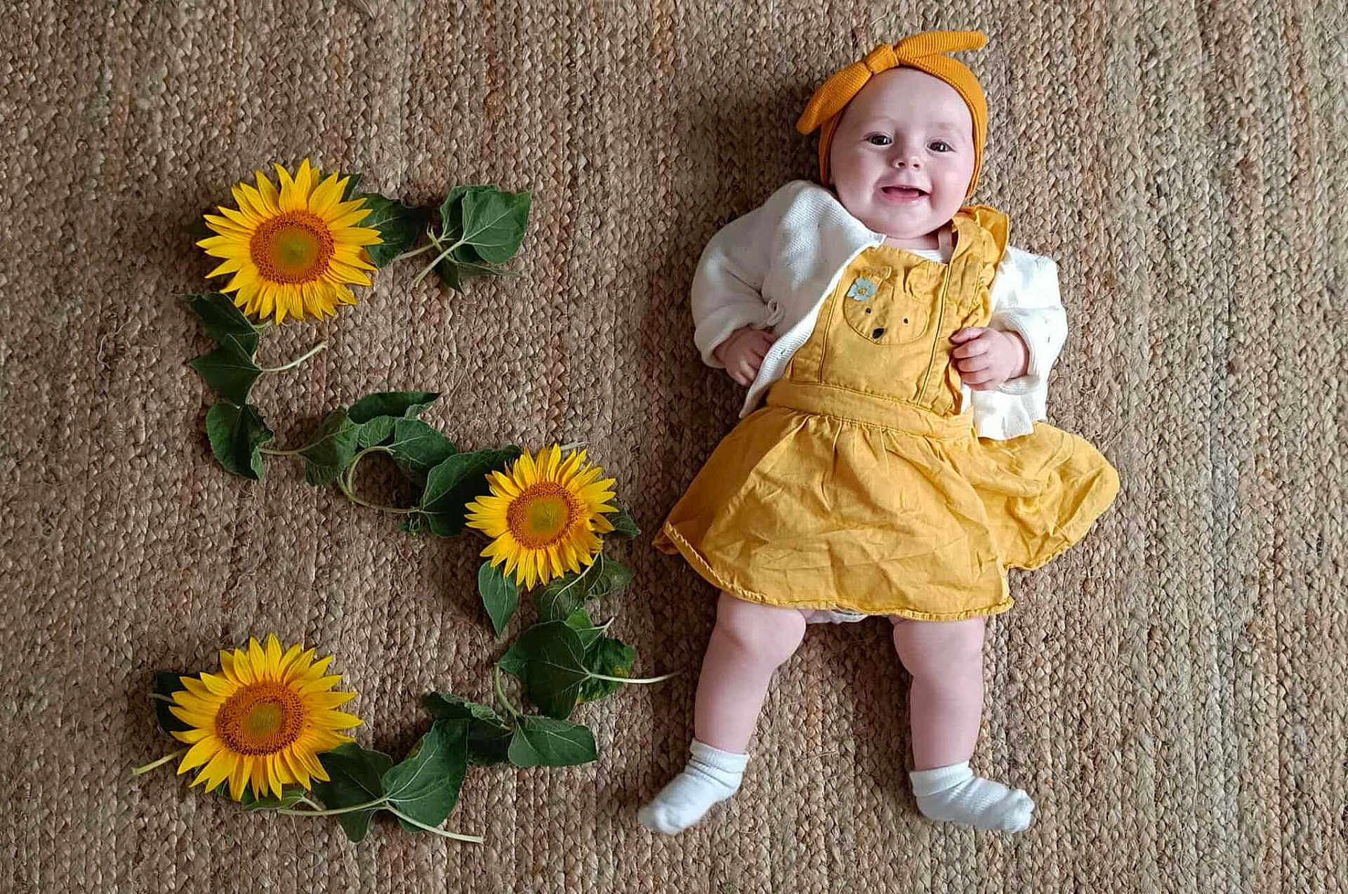 Rosalya participe au concours pour gagner de l'argent avec cette photo : baby, child, smiling, yellow_dress, headband, sunflowers, green_leaves, woven_rug, indoor, laying_down, happy, cute, infant, white_socks, natural_light, portrait, flower_arrangement, celebration, number_three, skin