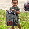 Isaiah is registered to the contest to win money with this photo: child, toddler, smiling, outdoor, halloween, costume, yard_sign, ghost, pumpkin, black_cat, spooky, grass, street, cars, parking, camouflage_shirt, shorts, sandals, portrait, daylight