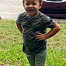 child, toddler, portrait, outdoor, grass, lawn, parked_car, vehicle, camouflage_shirt, shorts, standing, hands_on_hips, curly_hair, smiling, facial_expression, daylight, residential, front_yard, photography, person
