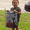 bat, camouflage_shirt, cars, child, costume, curly_hair, ghost, grass, halloween, lawn, outdoor, portrait, pumpkin, sandals, smiling, standing, street, toddler, treat_bag, witch
