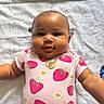 arms, baby, blanket, child, close_up, eyes, face, flower_pattern, fruit, indoor, infant, lying_down, necklace, photography, pink_outfit, portrait, round_cheeks, smiling, strawberry_pattern, white_background