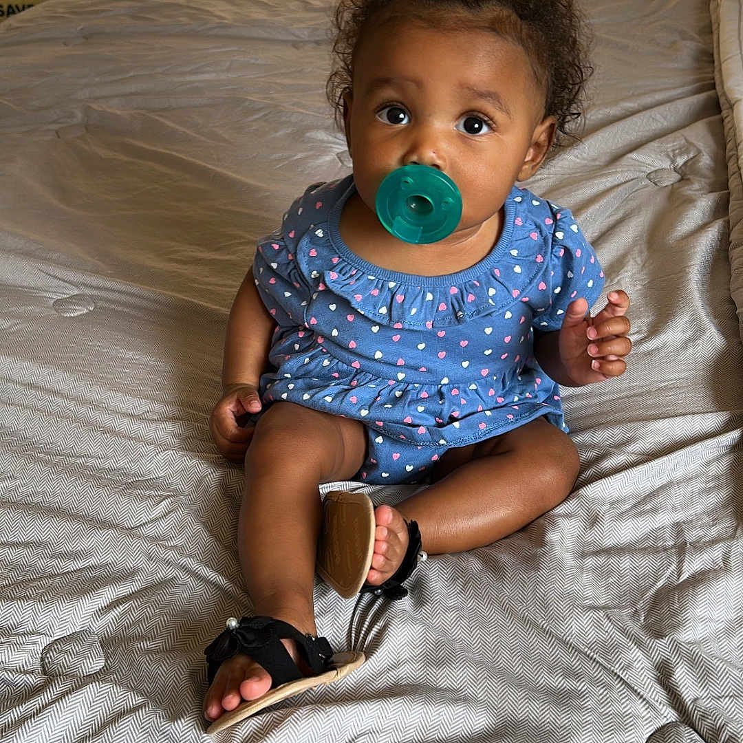Ariana is registered to the contest to win money with this photo: baby, child, pacifier, blue_dress, curly_hair, indoor, bed, blanket, sunlight, curtains, footwear, sitting, looking, young_child, cute, infant, person, soft_light, home, casual