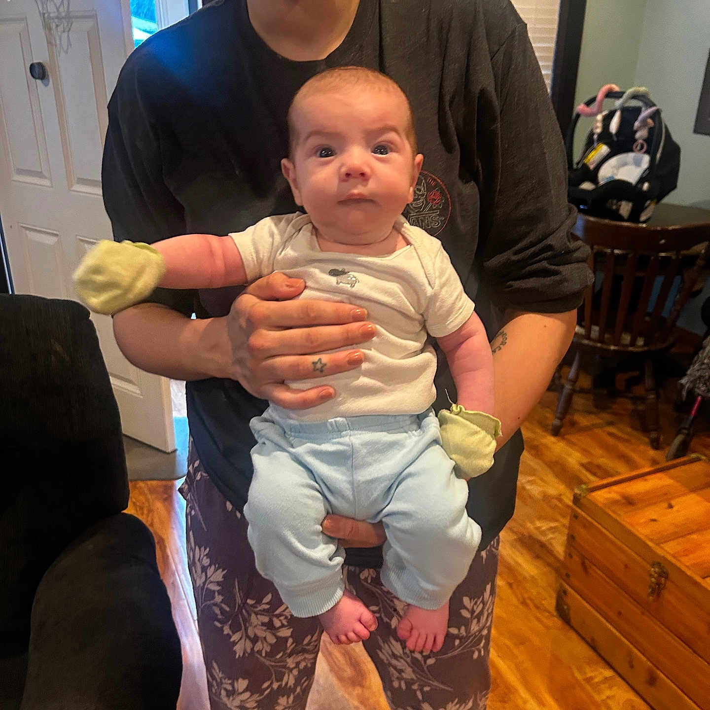 Emilio is registered to the contest to win money with this photo: baby, car_seat, chair, clothing, door, expression, face, floor, furniture, hand, holding, home, indoor, mittens, pants, person, table, tattoo, window, wooden_floor