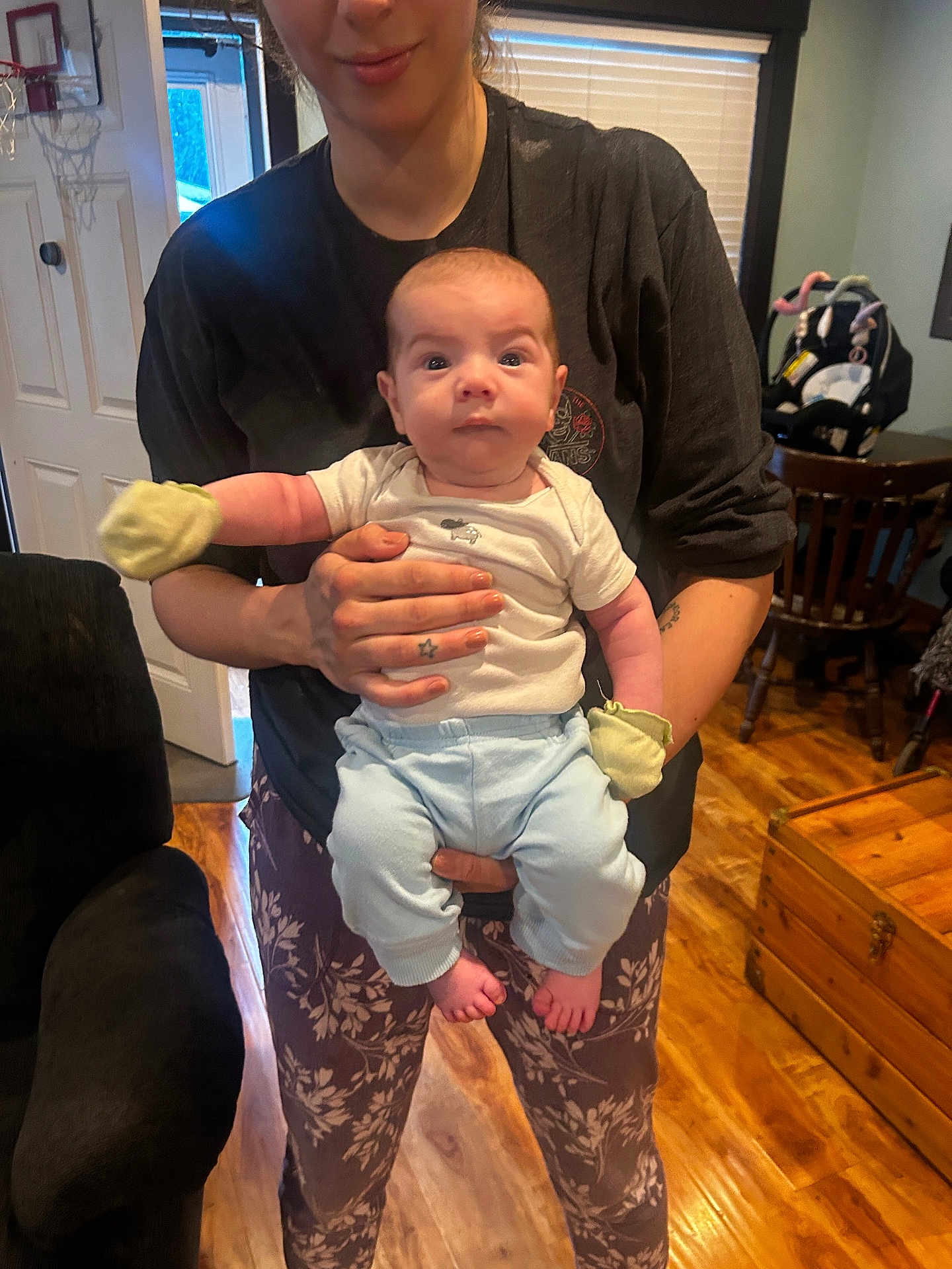 Emilio is registered to the contest to win money with this photo: baby, person, indoor, clothing, mittens, pants, floor, wooden_floor, furniture, chair, table, window, door, car_seat, tattoo, hand, face, expression, holding, home