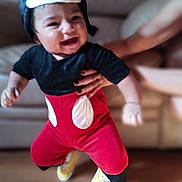 Andres is registered to the contest to win money with this photo: baby, child, smiling, costume, mickey_mouse, hat, red_pants, yellow_shoes, indoor, living_room, couch, wooden_floor, person, cute, happy, playful, infant, hand, holding, blurred_background