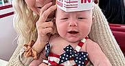 Zhelda joined the competition — help win amazing prizes! baby, woman, headband, hat, burger_box, indoor, restaurant, smile, happy, people, clothing, table, patriotic_outfit, baby_outfit, hand, face, person, holding, background, casual