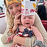 baby, woman, headband, hat, burger_box, indoor, restaurant, smile, happy, people, clothing, table, patriotic_outfit, baby_outfit, hand, face, person, holding, background, casual