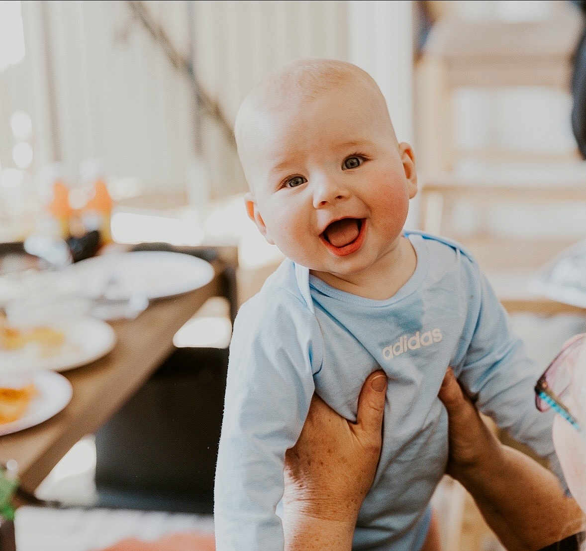 River joined the competition — help win amazing prizes! baby, infant, smiling, portrait, indoor, hands, adult_hands, eyes, mouth, blue_shirt, adidas, clothing, dining_table, plate, food, blurred_background, happy, family, cute, holding